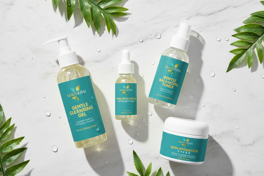 "Four Maudeish skincare products arranged in a flatlay: Gentle Hydrating Gel Cleanser, Gentle Balancing Toner, Dark Spot Serum for Sensitive Skin, and Hyaluronic Acid Hydration Cream on a white marble surface with green botanical leaves."